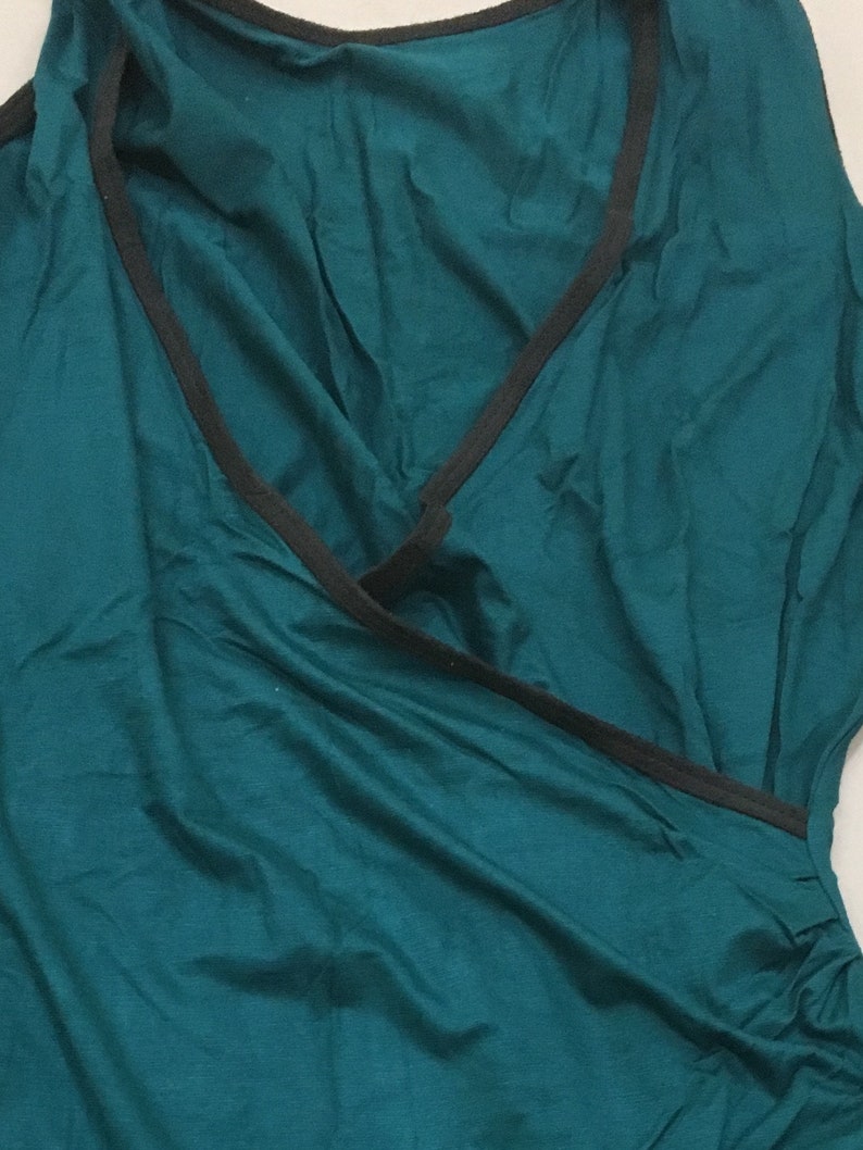 May include: A teal-colored sleeveless top with a wrap-style design. The neckline and edges are trimmed with a thin black band. The fabric has a soft, flowing appearance.