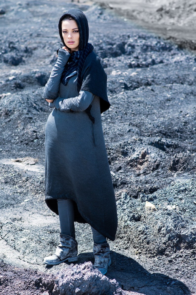 Puede incluir: Una persona con un vestido con capucha gris oscuro, bufanda y botas. El vestido tiene mangas largas y una falda fluida. Las botas son de color gris desgastado. El fondo es un paisaje rocoso y gris.