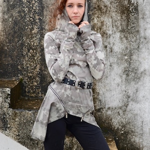 May include: A long-sleeved hooded tunic featuring a light gray and white digital camouflage pattern, thumbhole cuffs, and a diagonal silver-toned zipper detail at the hem. The garment is worn with a black belt featuring a silver buckle and black pants.