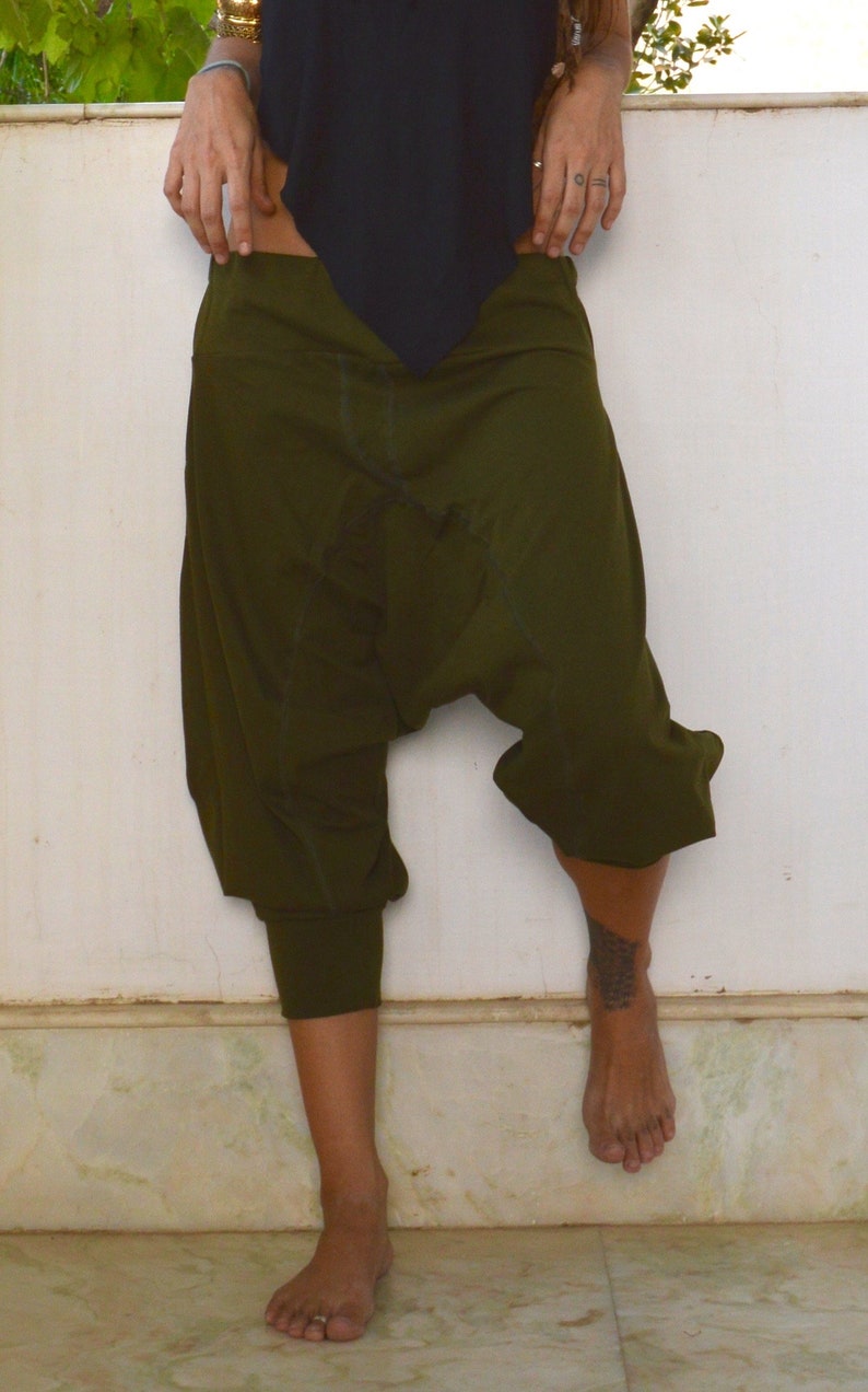 May include: Olive green harem pants with a relaxed fit. The pants have a gathered ankle cuff and a loose, baggy design. The model is wearing a black top. The pants are made of a soft, comfortable material.
