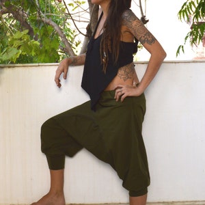 May include: A person wearing a black top and olive green harem pants. The pants have a relaxed fit with a gathered ankle. The top is sleeveless. The person is standing outside near a white wall and green foliage.