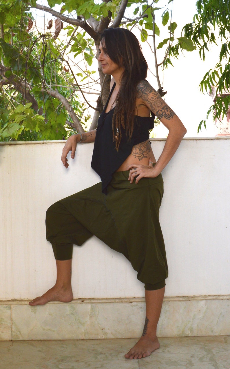 May include: A person wearing a black top and olive green harem pants. The pants have a relaxed fit with a gathered ankle. The top is sleeveless. The person is standing outside near a white wall and green foliage.