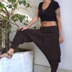 May include: A black crop top with a lace-up neckline and brown harem pants. The pants have a relaxed fit and cuffed ankles. The person is wearing a gold anklet. The background includes a white wall and a potted plant.