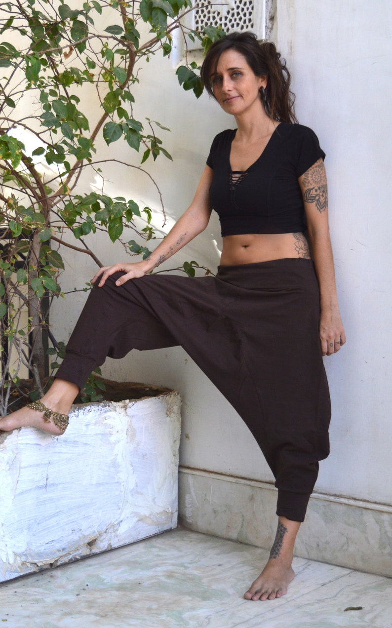 May include: A black crop top with a lace-up neckline and brown harem pants. The pants have a relaxed fit and cuffed ankles. The person is wearing a gold anklet. The background includes a white wall and a potted plant.