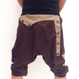May include: Brown and beige cotton trousers with a unique design featuring a flap with buttons and a side panel with buttons. The trousers have a loose fit and are cropped above the knee.