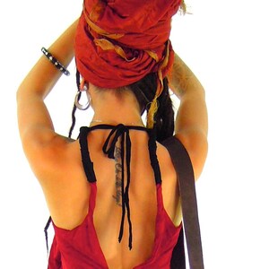 May include: A woman wearing a red and gold headscarf, a red halter top with black ties, and a black leather strap. She has dreadlocks and a tattoo on her back.