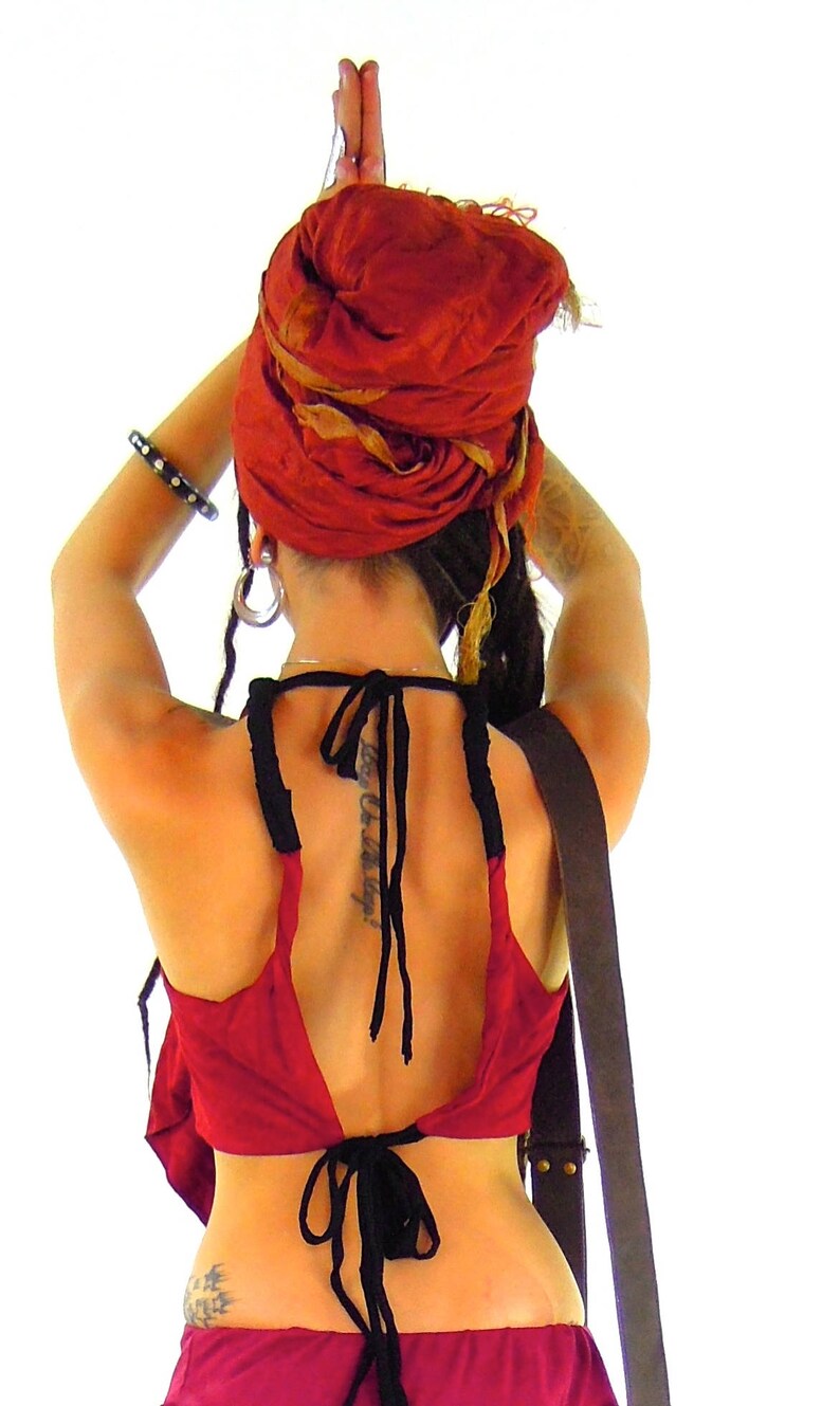 May include: A woman wearing a red and gold headscarf, a red halter top with black ties, and a black leather strap. She has dreadlocks and a tattoo on her back.