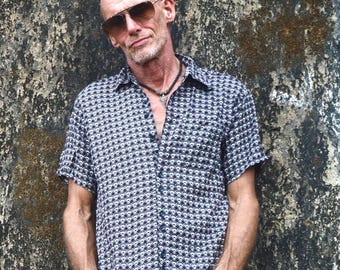 May include: A short-sleeved button-down shirt featuring a repeating geometric diamond pattern in black, white, and blue. The shirt has a classic collar and a full front button placket. It is worn with a dark cord necklace featuring a metallic pendant.