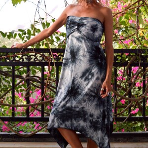May include: A strapless, knee-length dress with a tie-dye pattern in shades of grey and black. The dress has a gathered bodice and an asymmetrical hemline. The background features a black wrought iron railing and vibrant pink flowers.