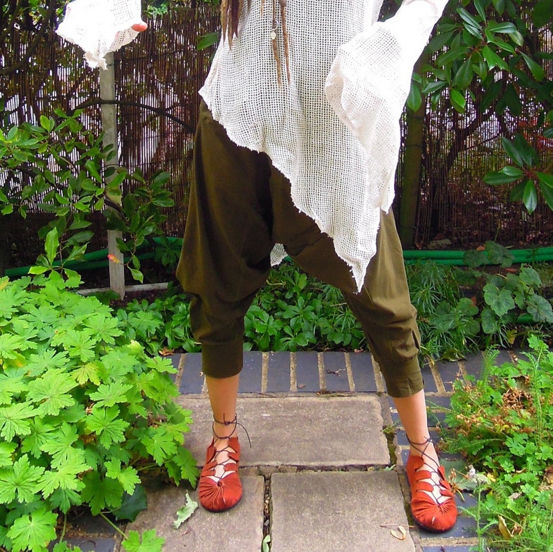 May include: Olive green harem pants and rust-colored lace-up shoes are featured in this image. The outfit is paired with a white, open-weave top. The setting is outdoors, with a brick pathway and lush greenery in the background. The style is bohemian.
