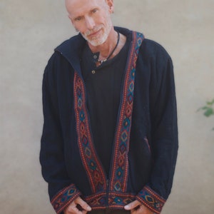 Puede incluir: Una chaqueta negra con capucha, cierre de cremallera y ribetes bordados de colores. La chaqueta tiene un corte holgado y un estilo informal.