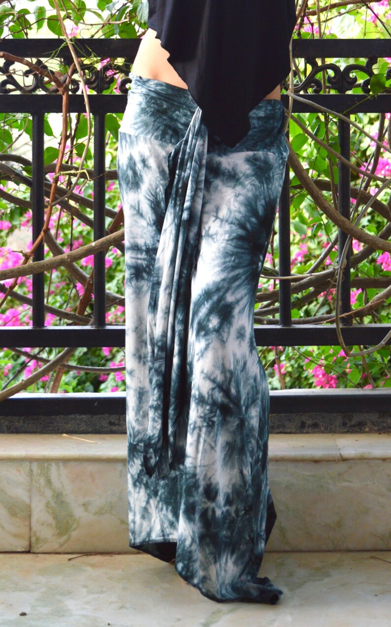 May include: Teal and white tie-dye trousers. The fabric is draped and knotted at the waist. The trousers are long and flowing, with a relaxed fit. The background includes a black metal railing and greenery.