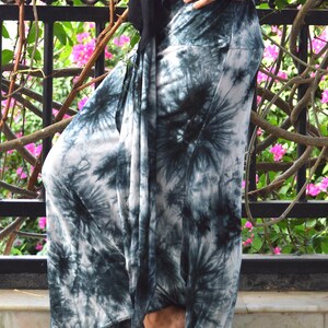 May include: White and teal tie-dye palazzo trousers. The wide-legged trousers have a high waist. A black top is visible. The background features a black metal railing and pink flowers.