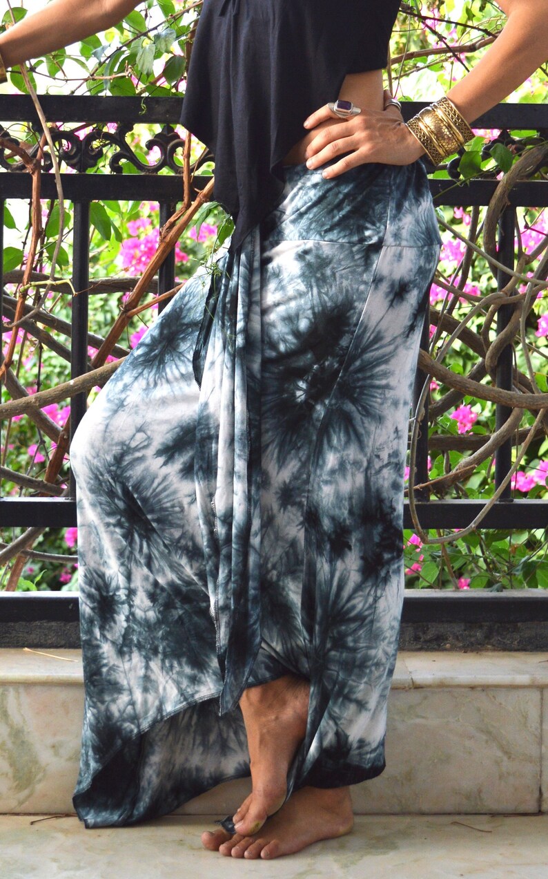 May include: White and teal tie-dye palazzo trousers. The wide-legged trousers have a high waist. A black top is visible. The background features a black metal railing and pink flowers.