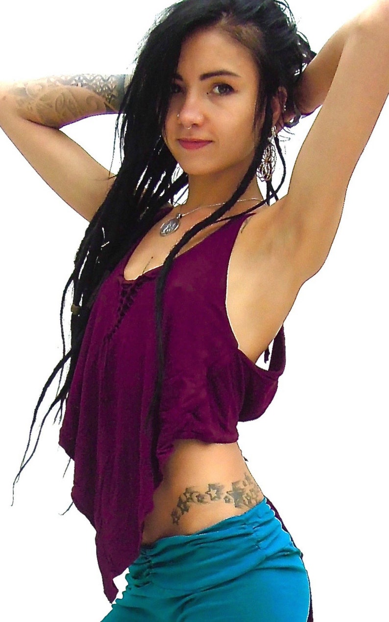 May include: A woman with long black dreadlocks is wearing a burgundy sleeveless top with a lace-up detail at the front. She is also wearing a turquoise skirt and has a star tattoo on her hip.