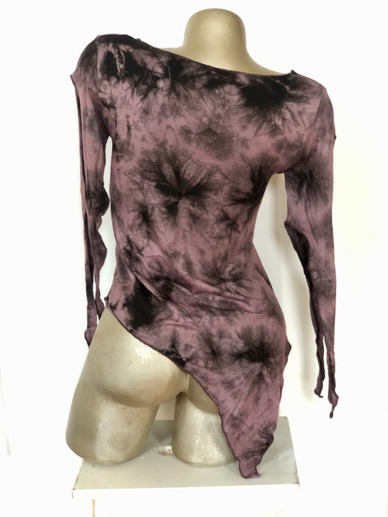 May include: A mauve and black tie-dye long-sleeve top with an asymmetrical hemline. The top has a one-shoulder design and is displayed on a mannequin. The fabric appears lightweight and flowing, with a dark, swirling pattern.
