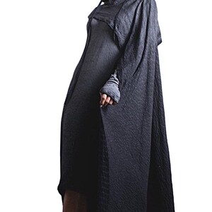 Puede incluir: Un vestido fluido gris oscuro con una capa a juego. El vestido tiene mangas largas y una tela texturizada. La modelo lleva botines negros con detalles de cadena. El fondo es blanco liso.
