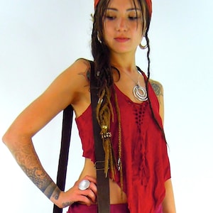 May include: A woman wearing a red top with a brown leather strap, a red bandana, and a silver necklace with multiple rings. She has long brown dreadlocks and is wearing a silver ring on her finger.