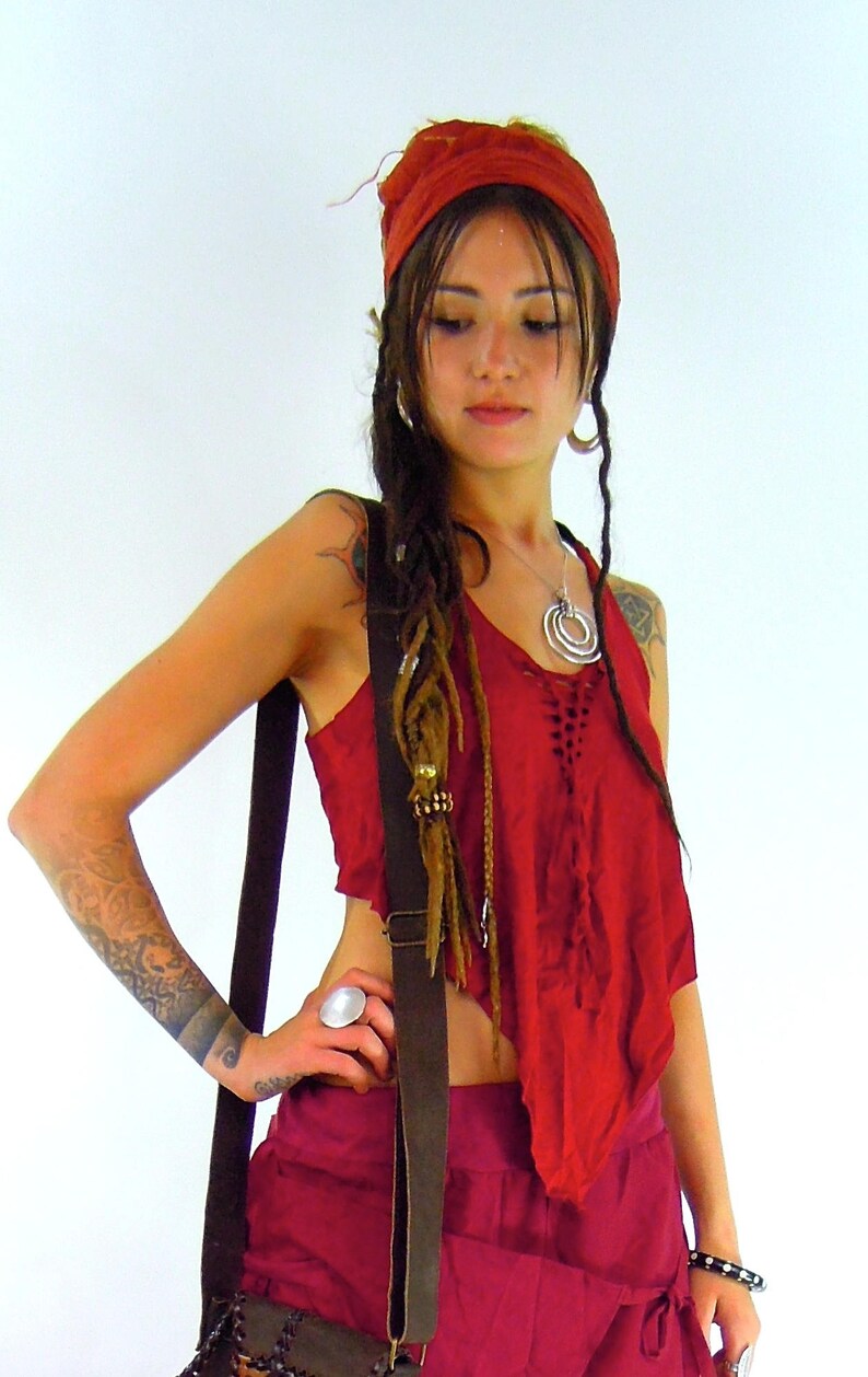 May include: A woman wearing a red top with a brown leather strap, a red bandana, and a silver necklace with multiple rings. She has long brown dreadlocks and is wearing a silver ring on her finger.
