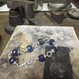 May include: A handmade bracelet featuring dark blue and teal-colored beads strung on a silver chain. The beads are interspersed with silver accents. The bracelet is displayed on a jeweler's workbench, surrounded by tools.