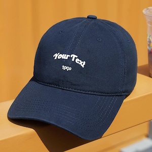 May include: A dark blue baseball cap with a white embroidered logo that says "Your Text Logo".