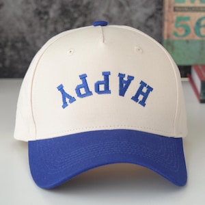 Könnte beinhalten: Eine weiße Baseballkappe mit einem blauen Schirm. Auf der Vorderseite der Kappe ist das Wort "HAPPY" in Blau gestickt.