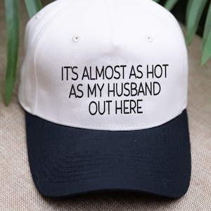 May include: A white and navy blue baseball cap with the text "IT'S ALMOST AS HOT AS MY HUSBAND OUT HERE" in black. The cap has a black button on top and a curved peak.