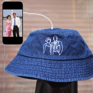 Può includere: Un cappello a secchiello in denim blu con un contorno ricamato bianco di una coppia. Il cappello ha una tesa classica ed è esposto con uno smartphone che mostra una foto della coppia. Il cappello è un accessorio elegante.
