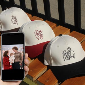 Può includere: Cappellini da baseball con ricami a tema familiare. I cappellini hanno una corona bianca e una visiera colorata in rosso o nero. Il ricamo raffigura una famiglia di tre persone. Un telefono mostra una foto di famiglia.