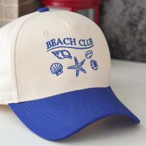 Puede incluir: Una gorra de béisbol blanca y azul con el texto "Beach Club" bordado en la parte delantera. La gorra tiene una visera azul y un diseño de conchas y una estrella de mar.
