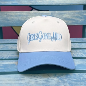 Puede incluir: Gorra de béisbol blanca y azul claro con el texto bordado "GIRLS GONE MILD" en escritura azul claro. La gorra tiene una visera curva y un botón en la parte superior. El sombrero está sobre una superficie de madera azul.