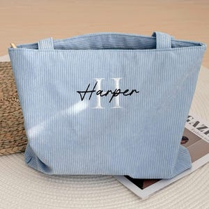 May include: A light blue corduroy tote bag with the name "Harper" embroidered in black script and white block letters. The bag has two handles and a rectangular shape. The bag is sitting on a white surface.