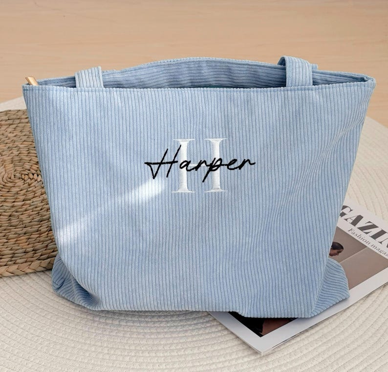 May include: A light blue corduroy tote bag with the name "Harper" embroidered in black script and white block letters. The bag has two handles and a rectangular shape. The bag is sitting on a white surface.