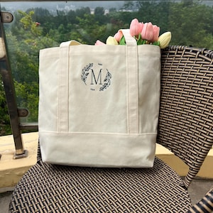 Op de afbeelding: Een witte canvas tote bag met een zwarte geborduurde krans en de letter "M" in het midden.