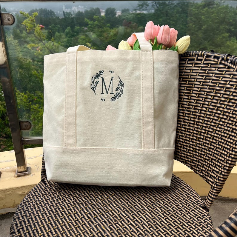 Op de afbeelding: Een witte canvas tote bag met een zwarte geborduurde krans en de letter "M" in het midden.