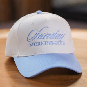 Könnte beinhalten: Weißes Baseball-Cap mit hellblauem Schirm und gesticktem Text "Sunday MORNING CLUB". Die Kappe hat eine runde Krone und einen kleinen Knopf oben.