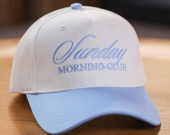 Pode incluir: Boné de beisebol branco com aba azul clara e texto bordado "Sunday MORNING CLUB". O boné tem uma coroa arredondada e um pequeno botão no topo.