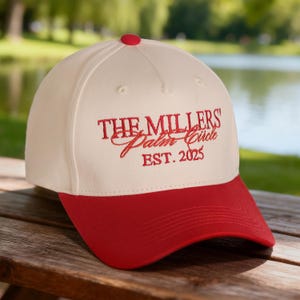 Könnte beinhalten: Cremefarbene Baseballkappe mit rotem Schirm und Knopf. Die Kappe hat den gestickten Text "THE MILLERS' Palm Circle EST. 2025" in roter Schrift. Der Hintergrund ist eine verschwommene Szene im Freien.