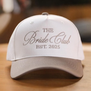 Puede incluir: Gorra de béisbol blanca con visera y botón superior grises. La gorra presenta el texto bordado "THE Bride Club EST. 2025" en fuente cursiva. El sombrero está hecho de una tela suave y probablemente sea para una despedida de soltera.