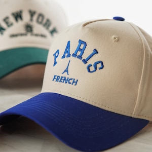 Op de afbeelding: Beige baseballcap met een blauwe klep, geborduurd met het woord "PARIS", een afbeelding van de Eiffeltoren en het woord "FRENCH". Een andere pet op de achtergrond is geborduurd met "NEW YORK".