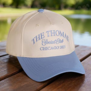 Op de afbeelding: Een baseballpet met een crèmekleurige kroon en een lichtblauwe klep. De tekst "THE THOMAS Social Club CHICAGO 2025" is lichtblauw op de voorkant geborduurd. De pet heeft een knoop bovenop.