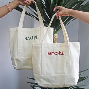 Op de afbeelding: Twee witte canvas tassen met handvatten. De tas aan de linkerkant heeft de naam "RACHEL" geborduurd in groen. De tas aan de rechterkant heeft de naam "BEYONC&Eacute;" geborduurd in rood.