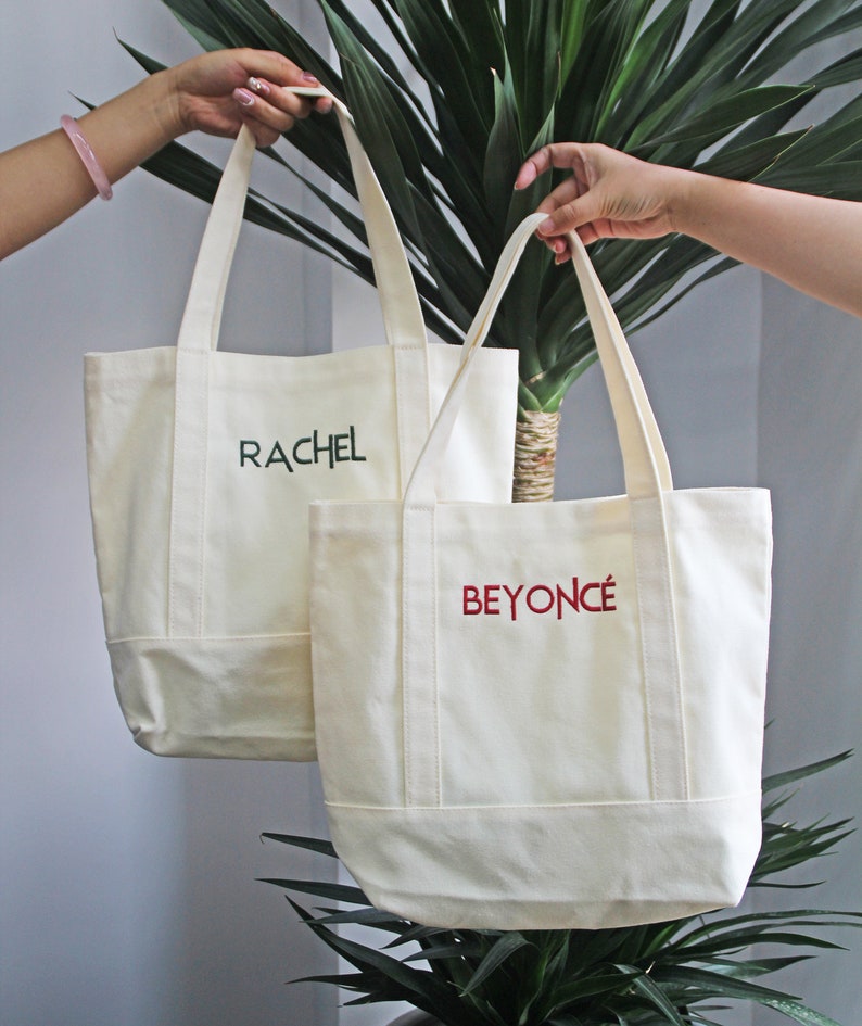 Op de afbeelding: Twee witte canvas tassen met handvatten. De tas aan de linkerkant heeft de naam "RACHEL" geborduurd in groen. De tas aan de rechterkant heeft de naam "BEYONC&Eacute;" geborduurd in rood.