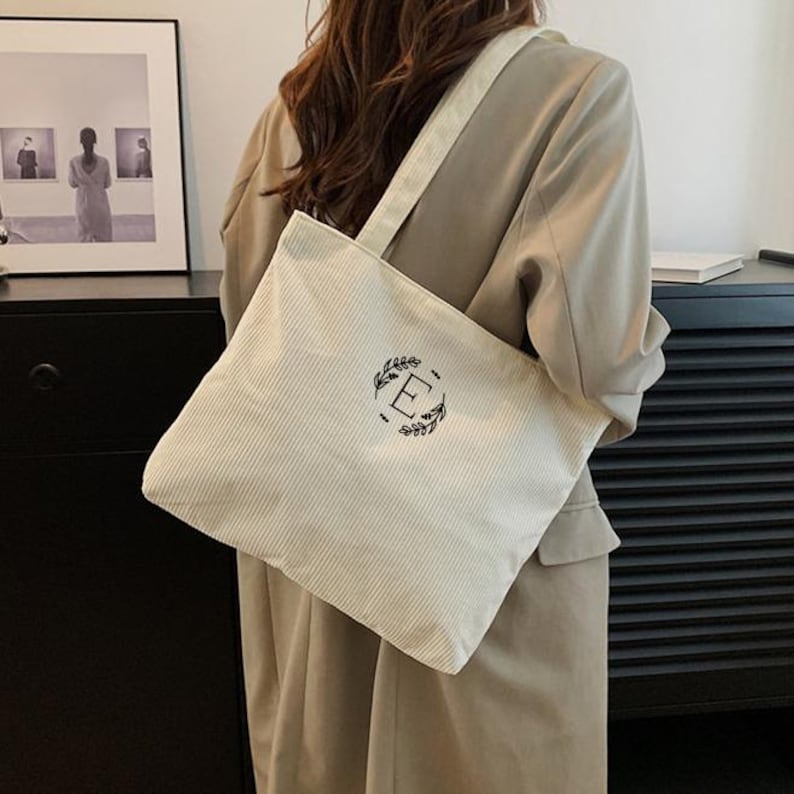 May include: Cream-colored corduroy tote bag with a shoulder strap. The bag features a black embroidered letter "E" surrounded by a leaf design. The bag is carried over the shoulder.