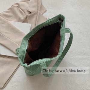 May include: Open view of a light green corduroy tote bag with a brown interior. The bag has two handles and a zipper closure. The text "The bag has a soft fabric lining" is visible.