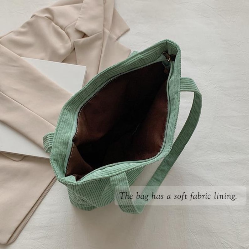 May include: Open view of a light green corduroy tote bag with a brown interior. The bag has two handles and a zipper closure. The text "The bag has a soft fabric lining" is visible.