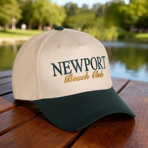 May include: A cream and dark green baseball cap with the embroidered text "NEWPORT Beach Club". The cap features a dark green peak and button, set against a blurred outdoor backdrop.