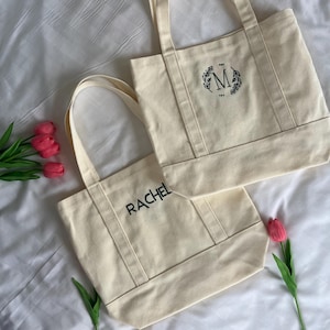 Op de afbeelding: Twee off-white canvas tassen met zwarte geborduurde letters. E&eacute;n tas zegt "RACHEL" en de andere heeft een krans met de letter "M" in het midden.