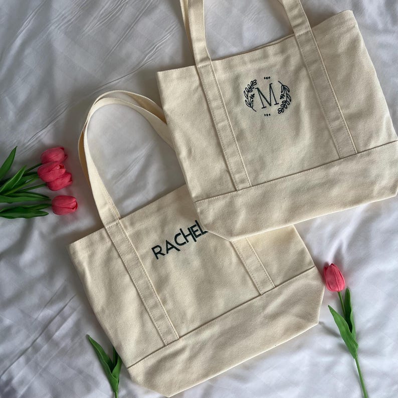 Op de afbeelding: Twee off-white canvas tassen met zwarte geborduurde letters. E&eacute;n tas zegt "RACHEL" en de andere heeft een krans met de letter "M" in het midden.