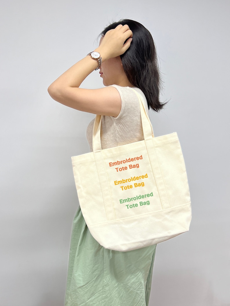 Op de afbeelding: Een witte canvas tote bag met de tekst "Embroidered Tote Bag" drie keer herhaald in verschillende kleuren: oranje, geel en groen.